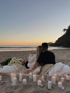 a man and woman sitting on top of a sandy beach next to the ocean with candles in front of them