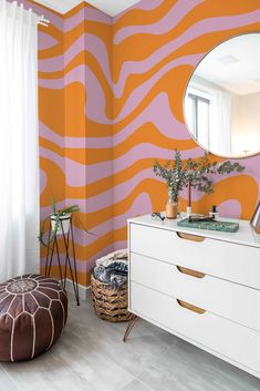 an orange and pink wall in a room with a white dresser, mirror and stool