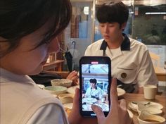 two people sitting at a table and one is looking at a cell phone while the other person stands in front of them