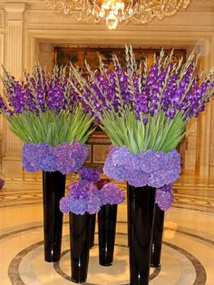 three black vases filled with purple flowers on top of a marble floor next to a chandelier