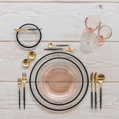 a table setting with black and gold plates, silverware and glasses on top of white wood planks