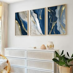 two paintings on the wall above a dresser in a room with white furniture and gold accents