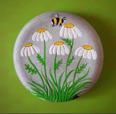 a painted rock with daisies and a bee on the top, sitting on a green surface