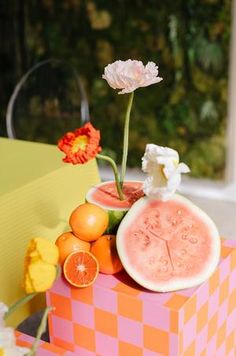 an arrangement of fruit and flowers in a vase on top of a pink checkered box