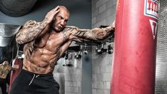 a shirtless man standing next to a red punching bag in a gym with tattoos on his arms