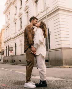 a man and woman kissing on the street in front of a white building with windows