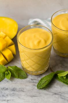 two glasses filled with mango custard next to sliced mangoes and mint leaves