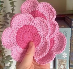 a hand is holding four pink crocheted coasters in front of bookshelves