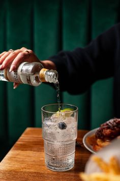 a person pouring water into a glass on top of a wooden table next to food