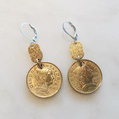 two gold coins are hanging from hooks on a white surface with blue beads and silver earwires