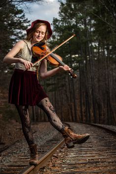 a woman in tights holding a violin on train tracks with trees and sky in the background