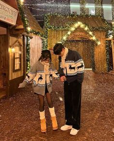 a man and woman standing next to each other in front of a wooden building with christmas lights