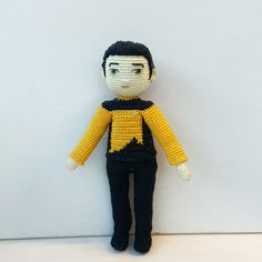 a crocheted doll is posed against a white wall, wearing a yellow and black outfit