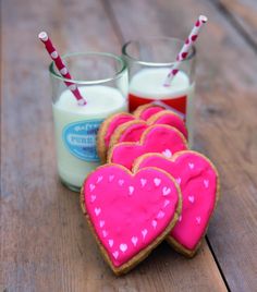 heart shaped cookies with pink icing next to two glasses of milk on a wooden table