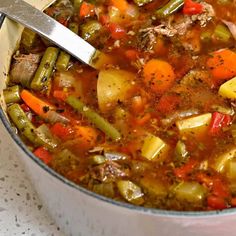 a pot filled with stew and vegetables on top of a white tablecloth next to a spoon