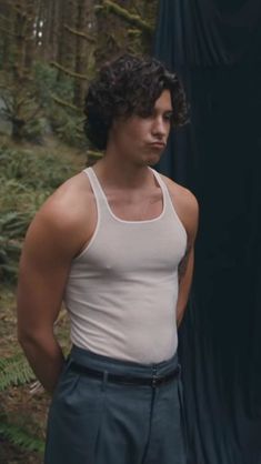 a man in white tank top standing next to a black curtain and looking at the camera