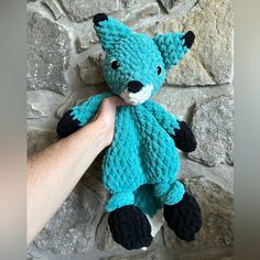 a hand holding a blue crocheted teddy bear on top of a stone wall