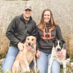 a man and woman sitting next to two dogs in front of a hay bale