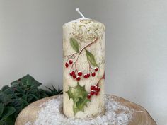 a white candle with holly and red berries on it sitting on top of a piece of wood