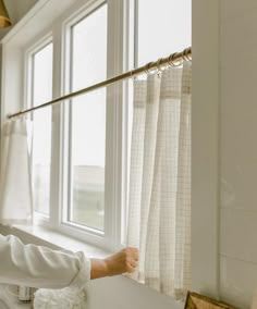 a woman is looking out the window with her hand on the curtain rod and she is wearing a white shirt