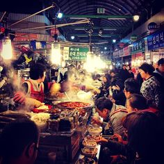 many people are eating food at an outdoor market area with lights on the ceiling and signs hanging above them