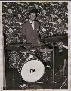 a black and white photo of a man playing the drums in front of a wall with stickers on it