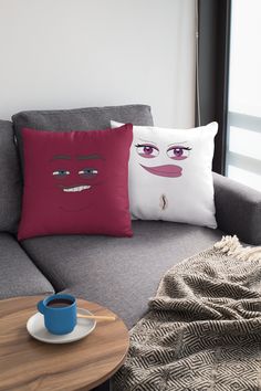 two pillows that have faces on them in front of a couch with a cup of coffee