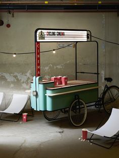 an old fashioned ice cream cart with red cups on it and two lounge chairs next to it