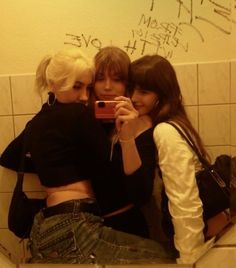 three young women taking a selfie in a bathroom stall with graffiti on the wall