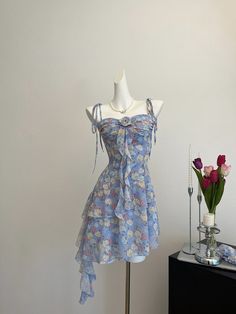 Circuitous and crafted with care, this blue floral dress weaves its way through timeless fashion with a modern twist. The layers of lightweight fabric create a gentle cascade, giving this dress a buoyant, ethereal quality that’s perfect for spring and summer events. Decorated with a vibrant array of floral patterns in soft hues, the dress features dainty shoulder straps and a rosette detail for a touch of romance. Its asymmetrical hemline and flowing silhouette ensure it moves gracefully with th Blue Ruffled Midi Dress For Summer, Summer Mini Dress For Garden Party In Blue, Blue Summer Mini Dress For Garden Party, Blue Floral Sundress For Summer, Blue Ruffled Midi Dress For Spring, Blue Sundress For Garden Party, Light Blue Floral Print Summer Dresses, Blue Floral Sundress For Garden Party, Light Blue Floral Print Sundress For Garden Party