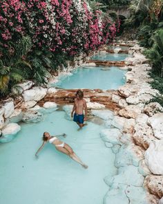 two people are sitting in the middle of a blue pool with rocks and flowers around them