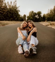 two young women sitting on the side of a road smiling at the camera, with their arms around each other