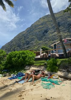 two people laying in the sand under a palm tree on a beach with mountains in the background