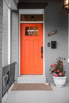 a bright orange front door on a gray house with flowers in the planter next to it