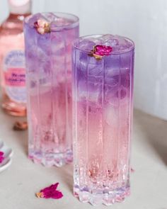 two pink and purple drinks sitting on top of a table next to bottles of alcohol