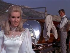 a woman in white dress standing next to an old fashioned car with man looking on