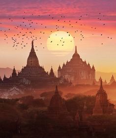 the sun is setting over an ancient city with many spires and birds flying in the sky