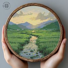 a hand holding up a cross stitch picture with water and mountains in the background,