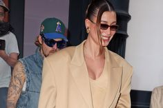 a woman wearing sunglasses and a tan blazer smiles as she walks past a man in a baseball cap