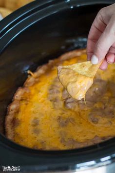 a hand holding a tortilla chip over a crock pot filled with cheese