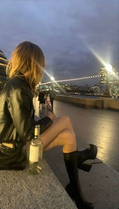 a woman sitting on the edge of a bridge with a bottle of wine in her hand