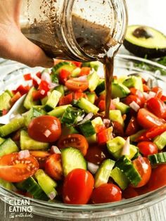 someone pouring dressing on a salad in a bowl with tomatoes, cucumbers and avocado