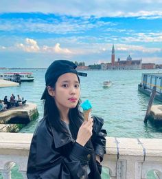 a woman is eating an ice cream cone near the water and boats in the background