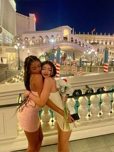 two young women hugging each other in front of a building at night with lights on