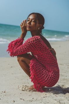 a woman sitting on top of a sandy beach next to the ocean wearing a red crocheted dress