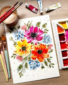 watercolor paints and brushes are sitting on a table next to an art card with flowers painted on it