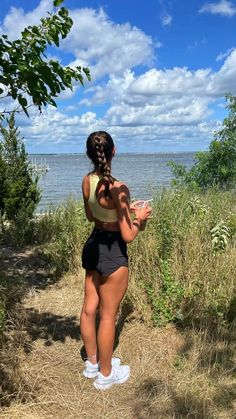 a woman standing on top of a grass covered field next to the ocean with her hands in her pockets