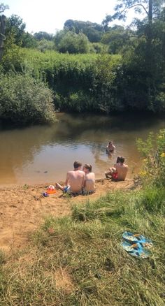 three people are sitting on the sand near a body of water with trees in the background