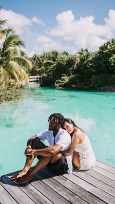 a man and woman sitting on a dock next to the water with palm trees in the background