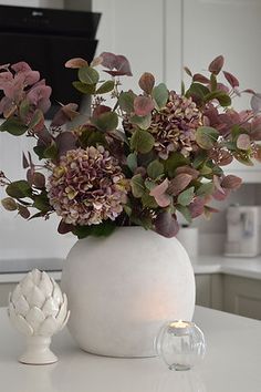 a white vase filled with purple flowers on top of a counter next to a candle